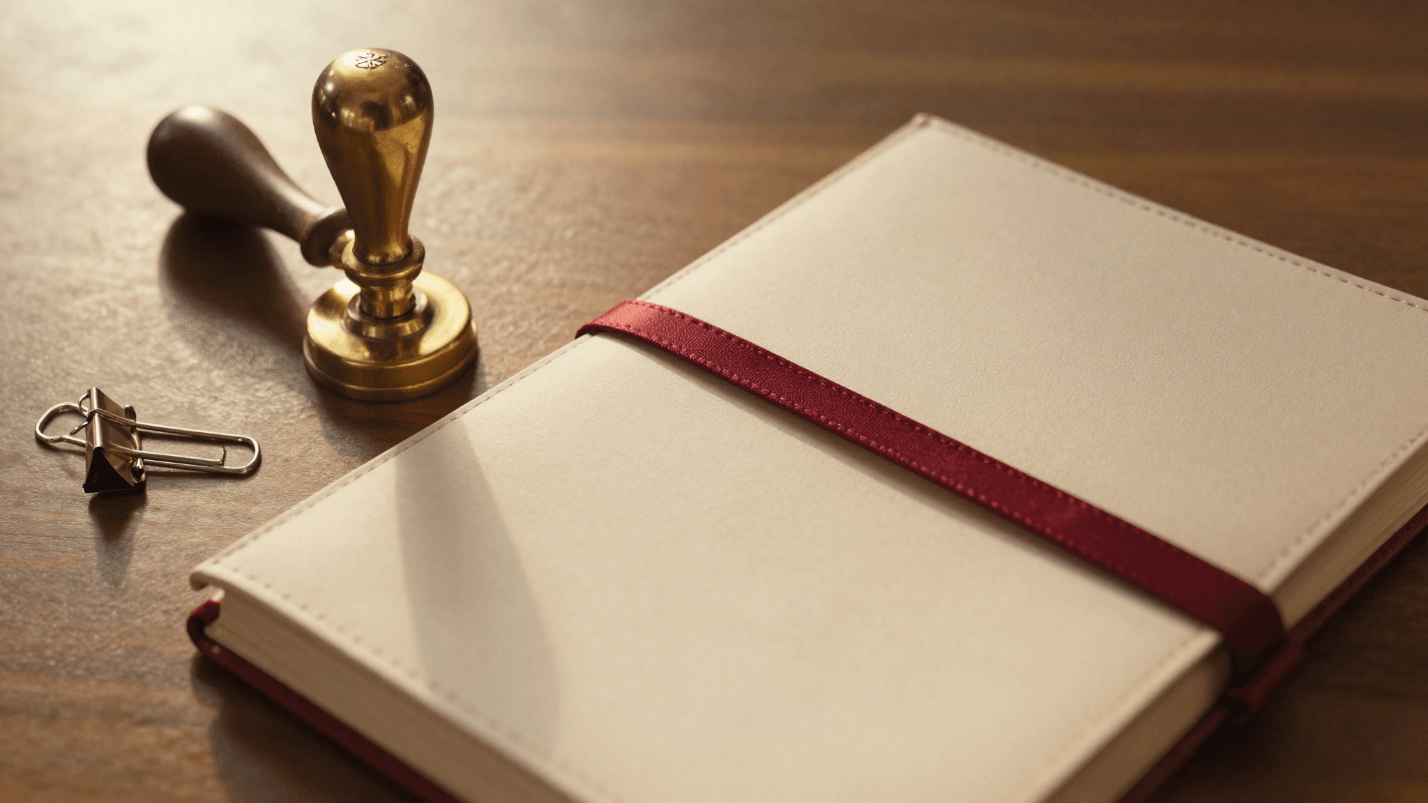 Top-down view of an ornate brass corporate seal embosser resting beside a closed leather portfolio tied with an oxblood ribbon, brass paper clip nearby, on a walnut desk in warm afternoon light